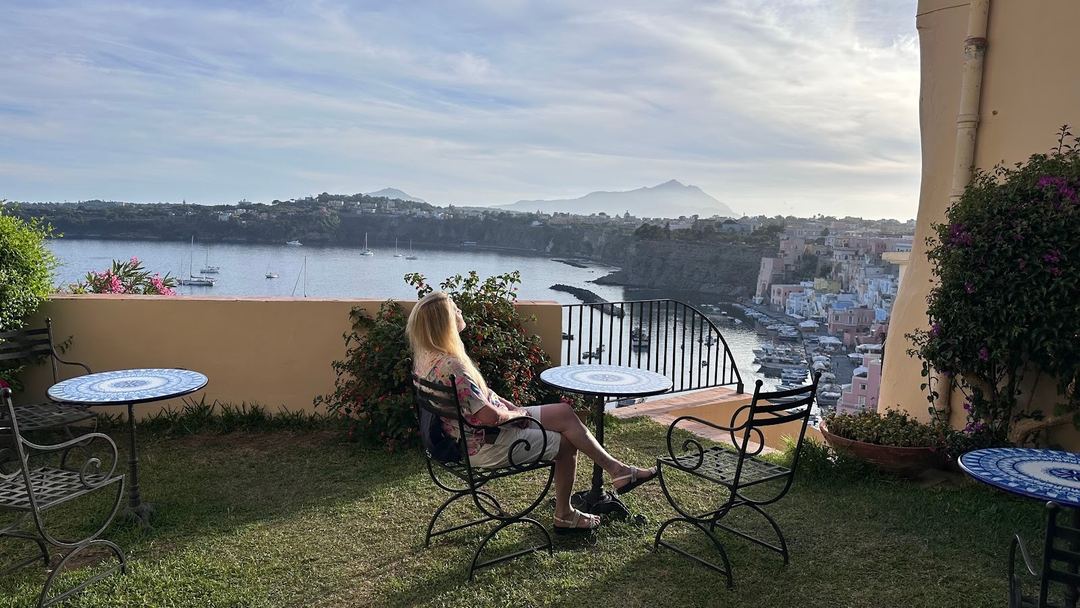 A guest enjoys the panoramic coastal view from the hotel's terrace overlooking a Mediterranean bay with sailboats and cliffside buildings.