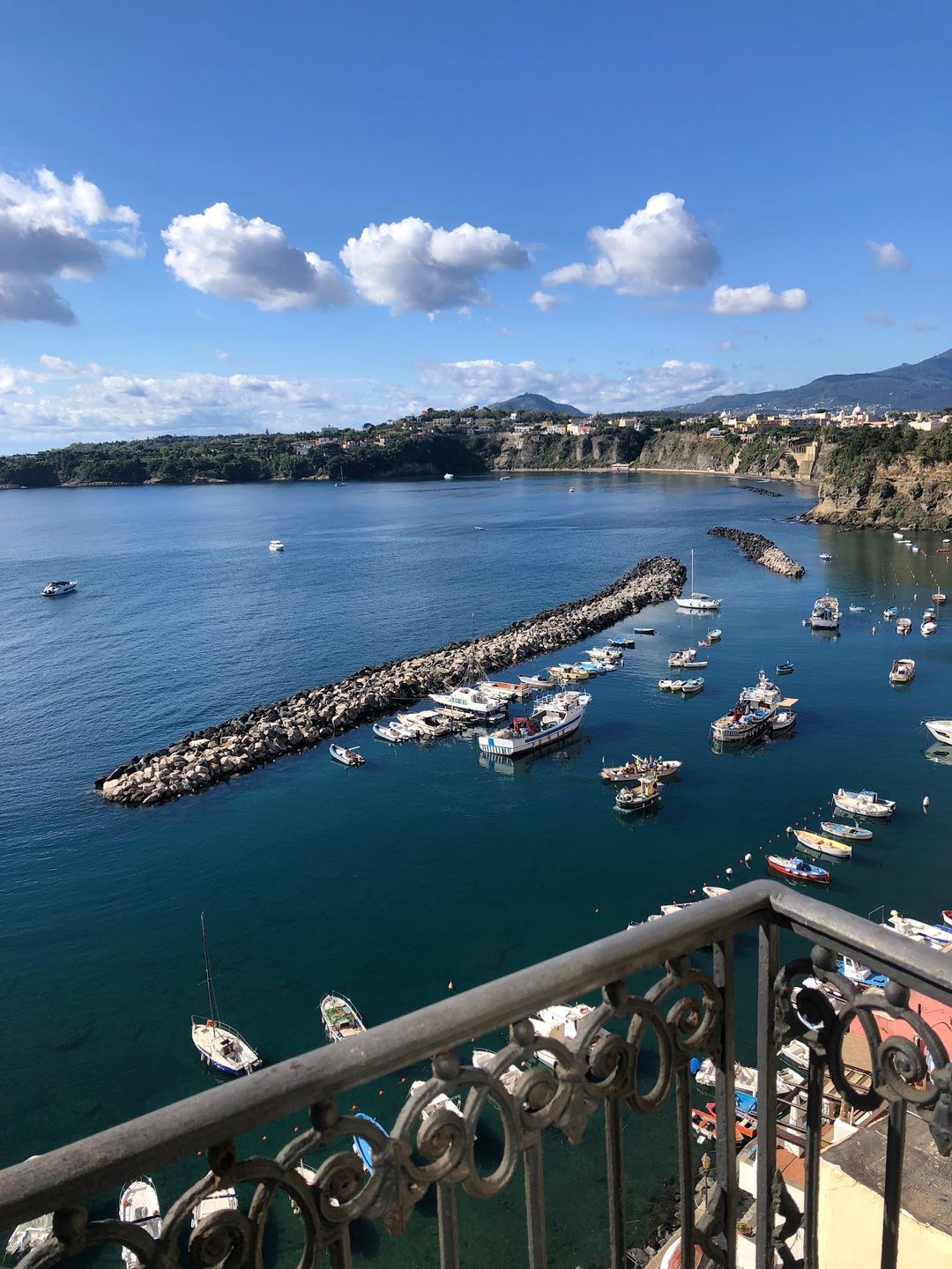 Panoramic view of a Mediterranean coastal harbor with moored fishing boats, a rocky breakwater, and hillside settlements under clear blue skies.