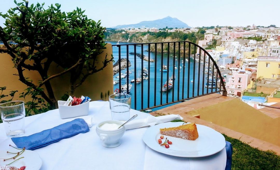 A seaside terrace dining setup overlooking a picturesque harbor with colorful buildings and mountains in the distance.