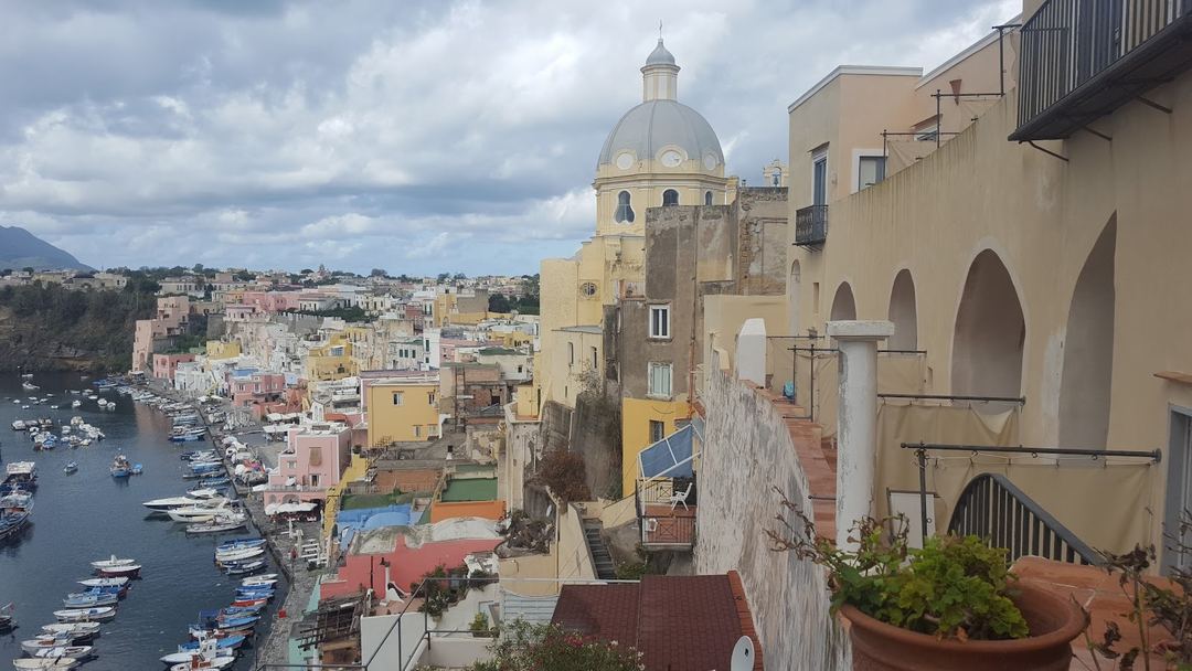 Panoramic view of Procida's colorful harbor village with the Church of the Assumption dome and Mediterranean Sea lined with fishing boats.