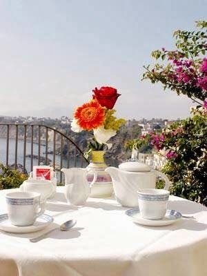 A seaside terrace table with fresh flowers and coffee service overlooking a coastal town and waterfront.